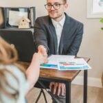 Real estate agent in suit shaking hands with client over property deal papers indoors.